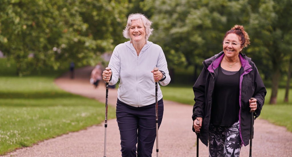 The image shows two older women walking along a path, each using poles.